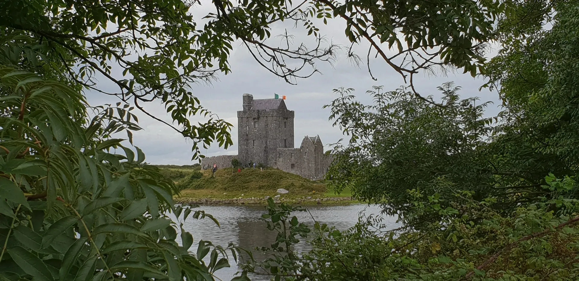 Stone tower across a river, framed by leafy green trees under a cloudy sky