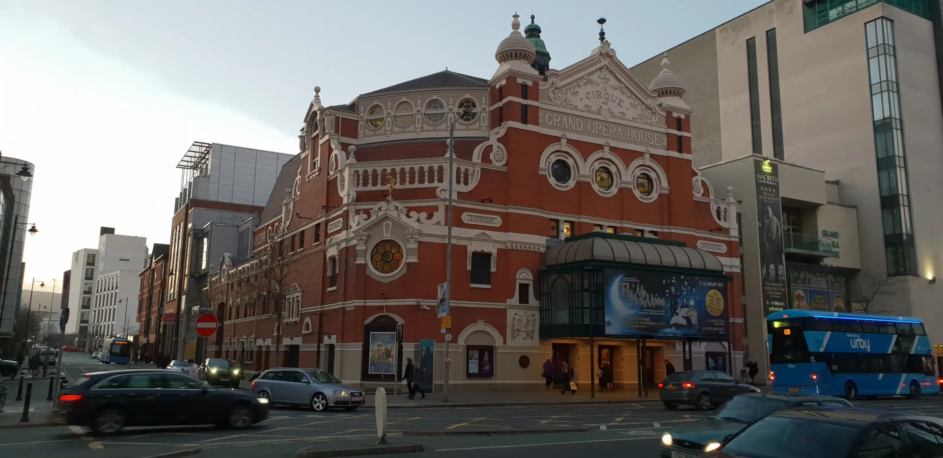 Red brick historic building with ornate white trim at a city intersection, cars and a blue bus passing by.