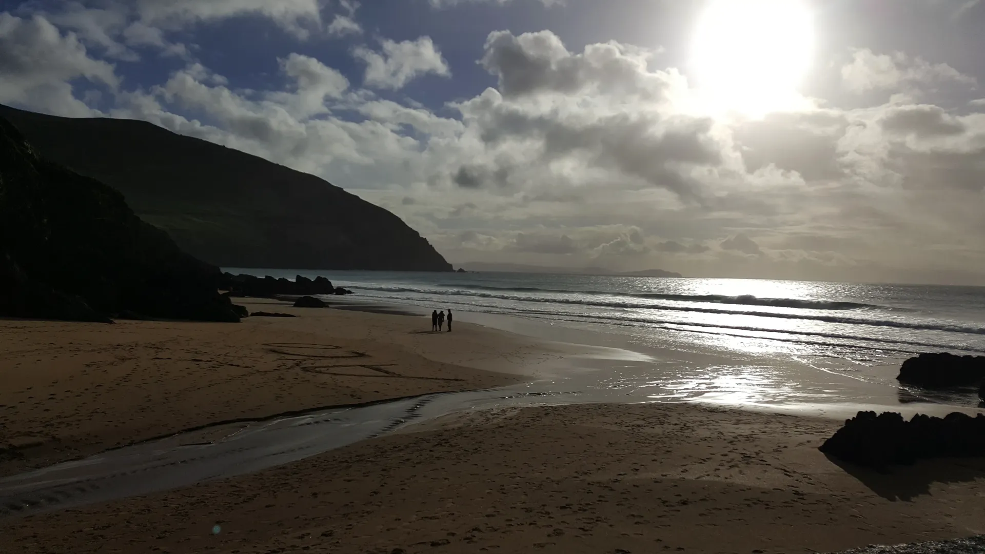 Sunlit beach at low tide with waves, rocky cliffs, and a few people in the distance