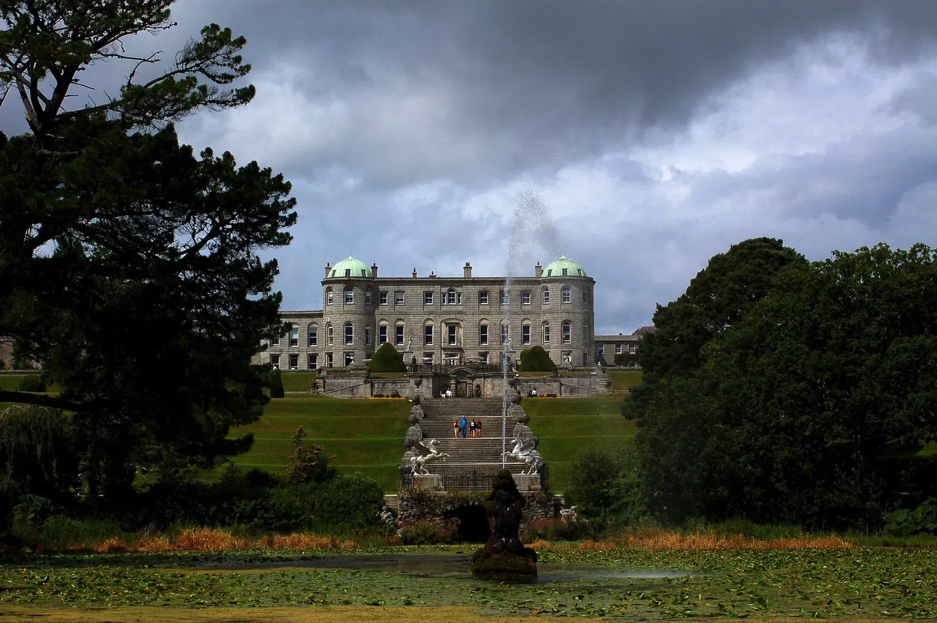 Grand historic mansion on a hill with stone steps, framed by trees under a cloudy sky