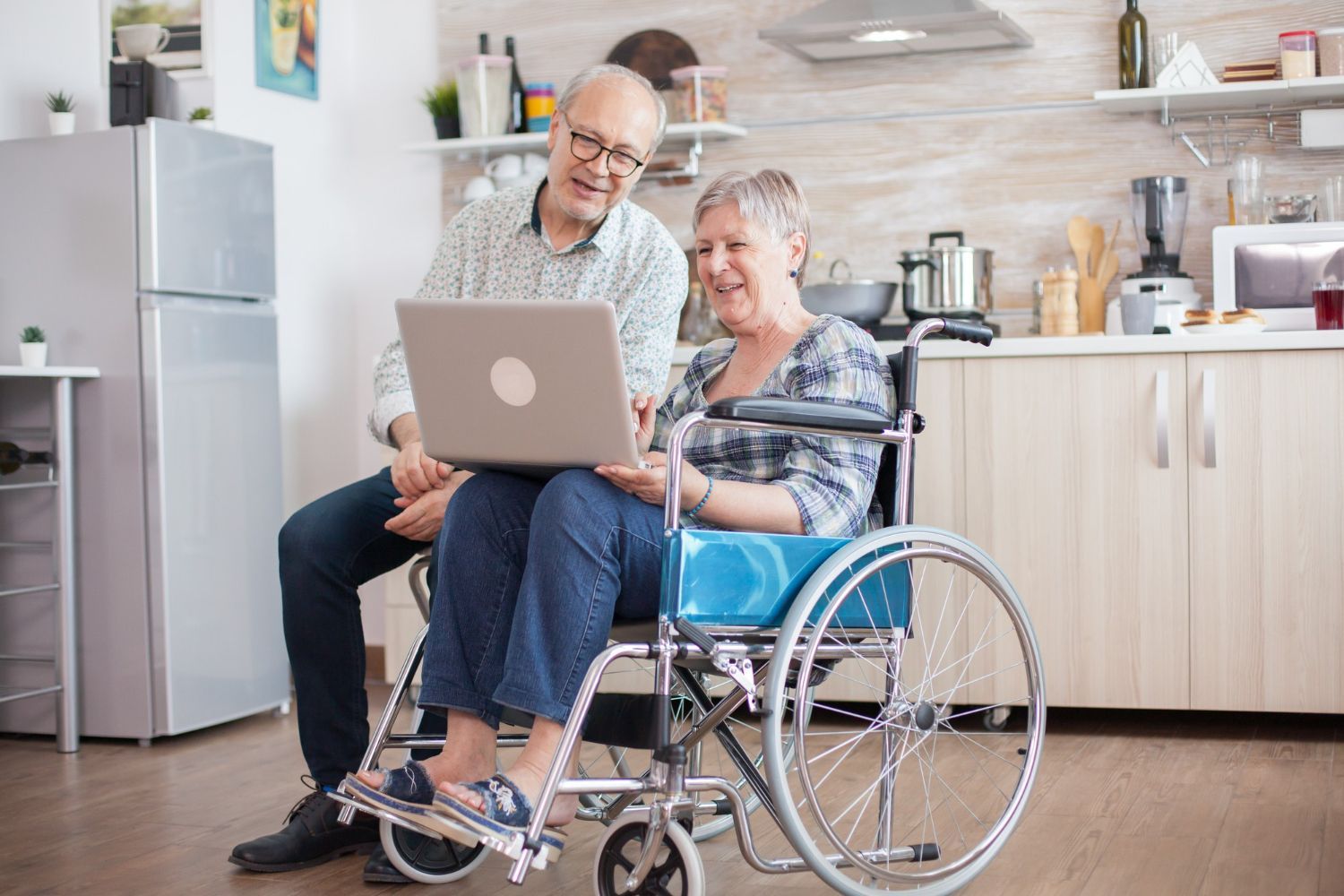 Older couple in kitchen: man looking at laptop with woman in wheelchair.