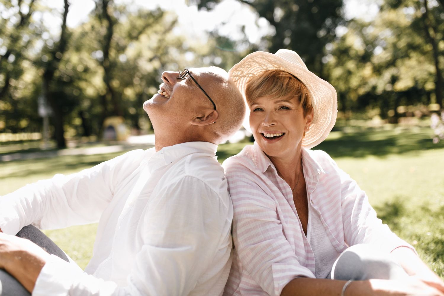 Mature couple smiles, back-to-back, on grass in sunny park. Man looks up, woman wears hat.