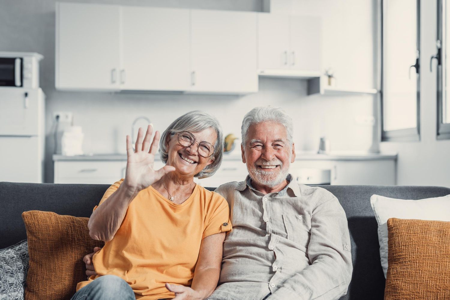 Smiling elderly couple on a couch, woman waving, sunny living room with kitchen.