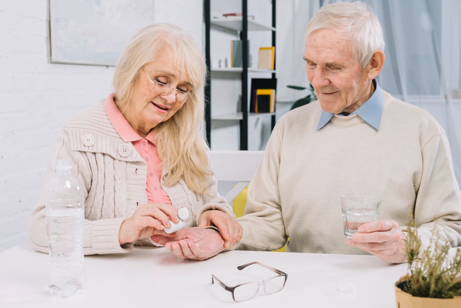 Elderly woman dispenses pills into her hand, man holds water glass. They sit at a table, glasses in foreground.