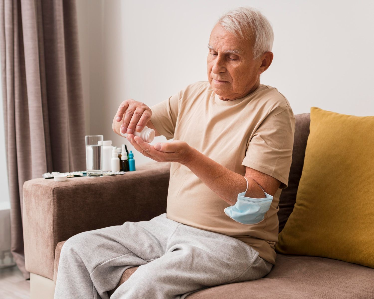 Elderly man on sofa, pouring pills into his hand. Face mask nearby.