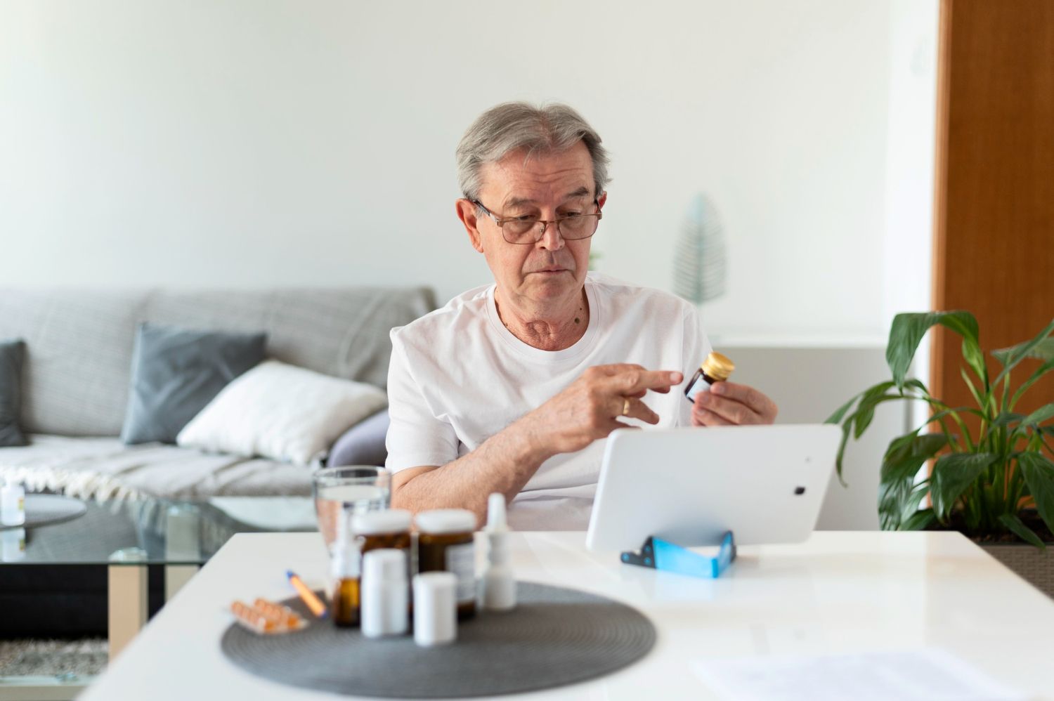 Elderly man consults a tablet while holding pills, sitting at a table with medications.