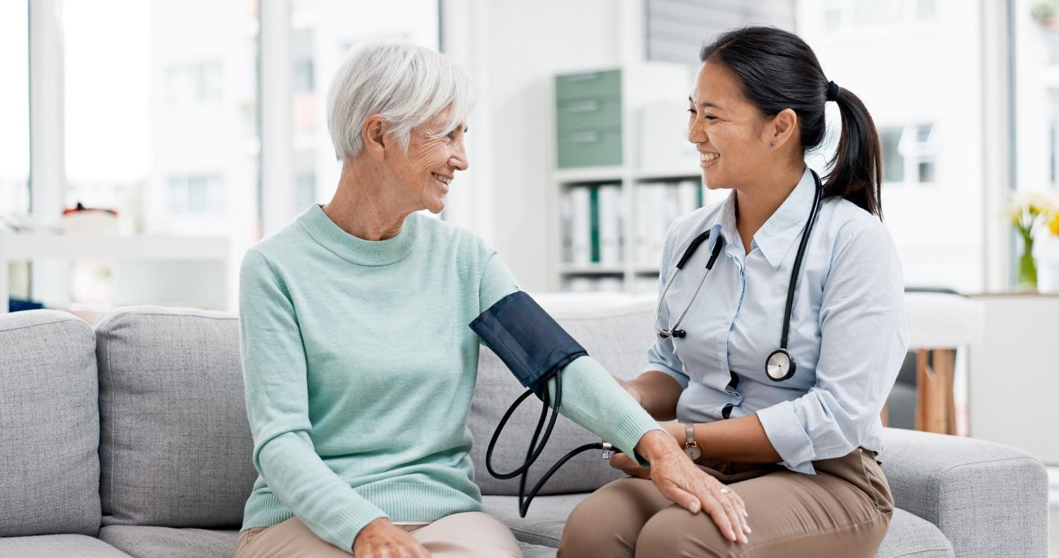 Doctor taking a senior woman's blood pressure on a couch. Both are smiling, indoors.