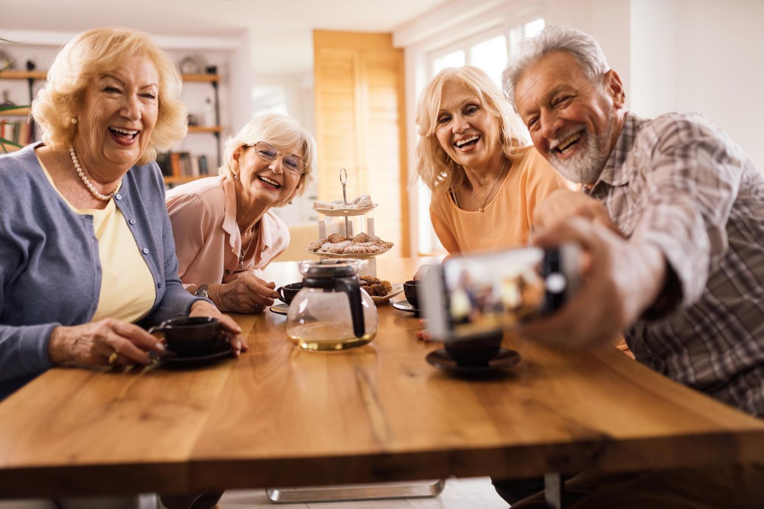 Four smiling seniors taking a selfie at a wooden table with coffee, light interior.
