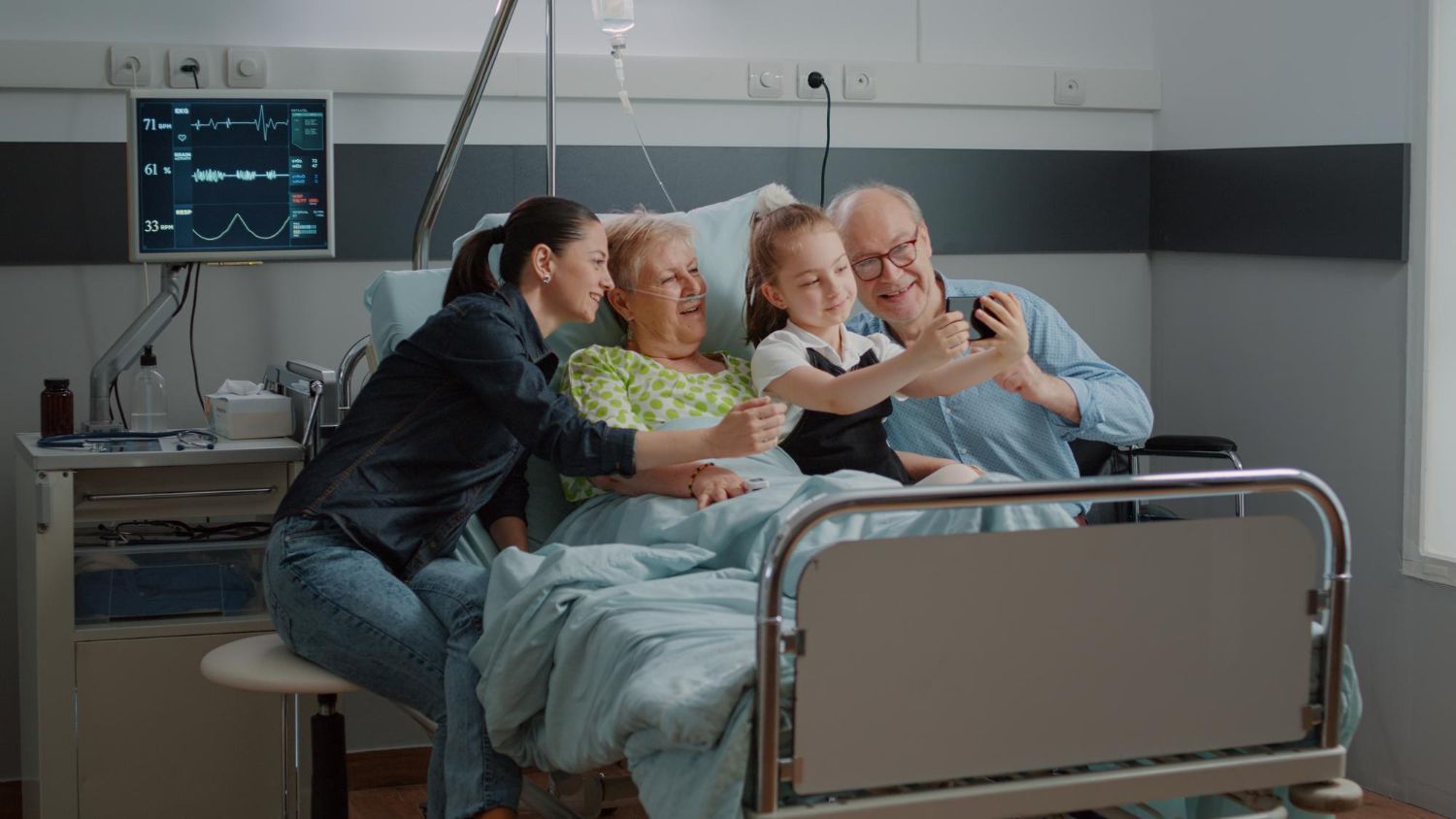 Family taking a selfie with elderly woman in hospital bed; daughter, granddaughter, and man smiling.