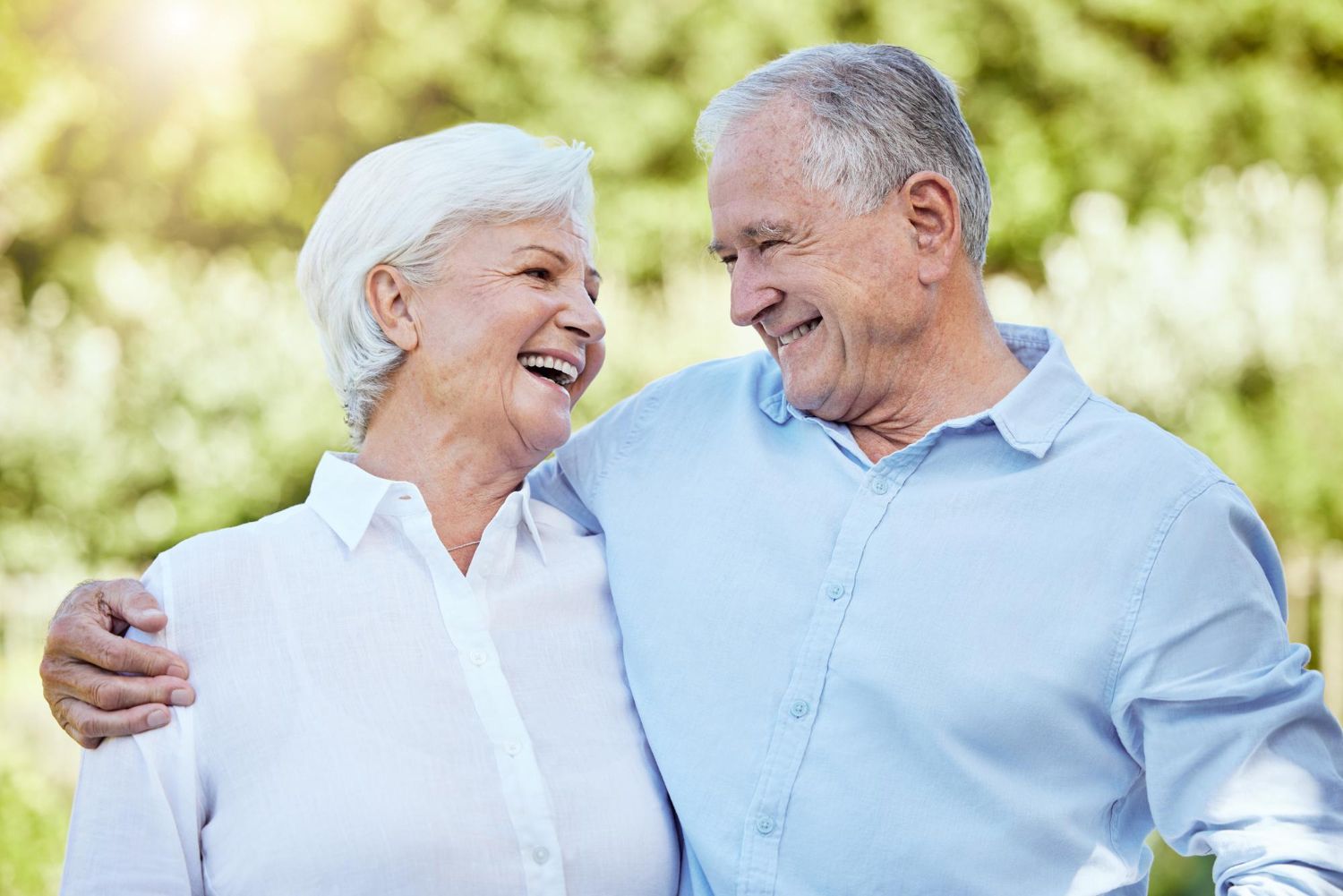 Smiling elderly couple outdoors, man's arm around woman, both looking at each other.