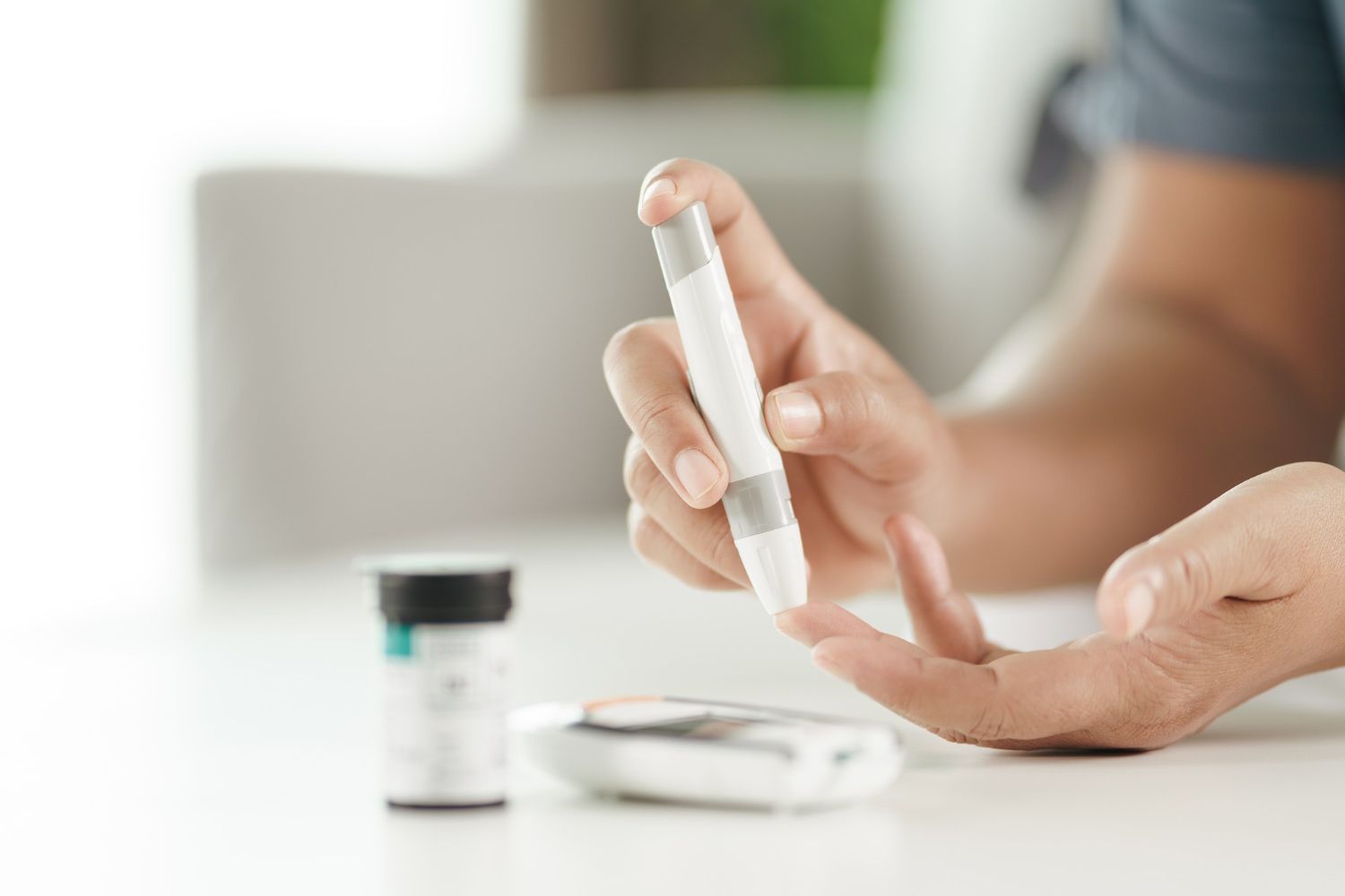 Person using a finger-prick device to test blood glucose levels on a white table next to a meter and vial.