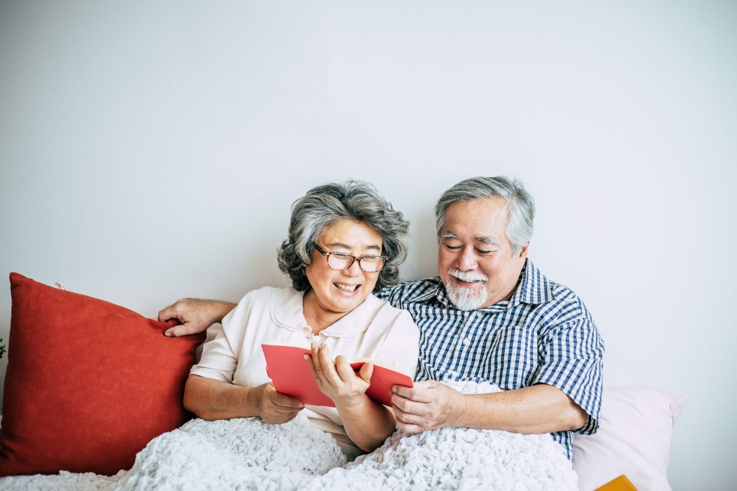 Elderly couple smiles while looking at a red book, sitting on a bed with red and pink pillows.