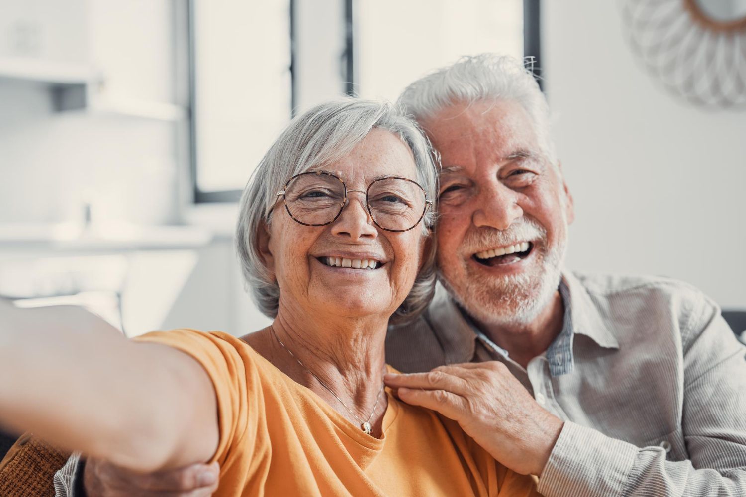 Smiling elderly couple taking a selfie indoors. The woman wears glasses and an orange shirt, the man a light blue shirt.