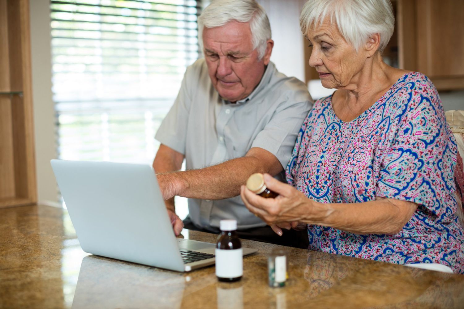 Elderly couple looking at laptop, holding medicine bottles, home interior.