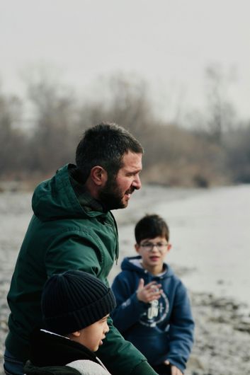 A man in a green jacket is talking to two boys on a beach.