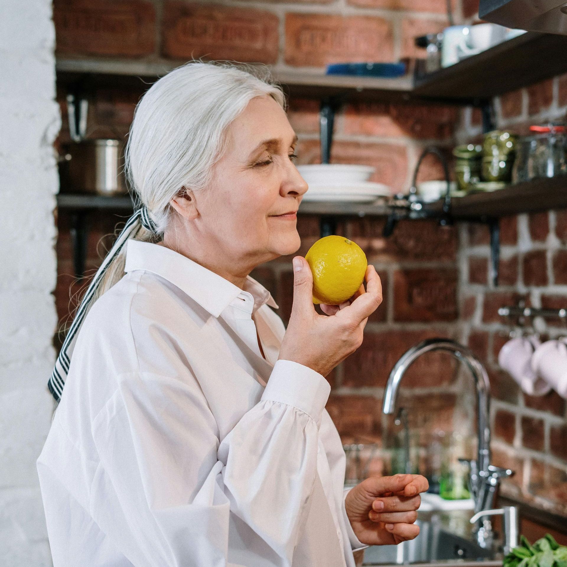 An elderly woman is holding a lemon in her hand in a kitchen.