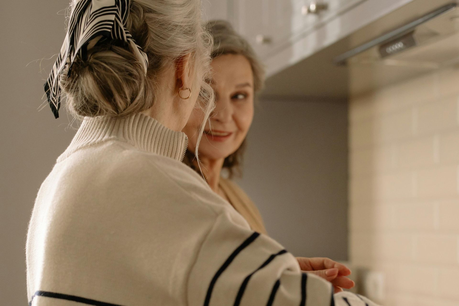 Two older women in a kitchen; one faces away wearing a headscarf, the other smiles warmly.
