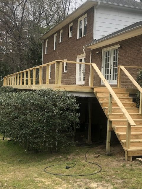 A newly constructed wooden deck and staircase attached to the side of a two-story brick house with trees in the background.
