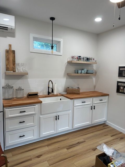A farmhouse kitchen with white cabinets, a white apron sink, wood countertops, floating wood shelves, and light floors.