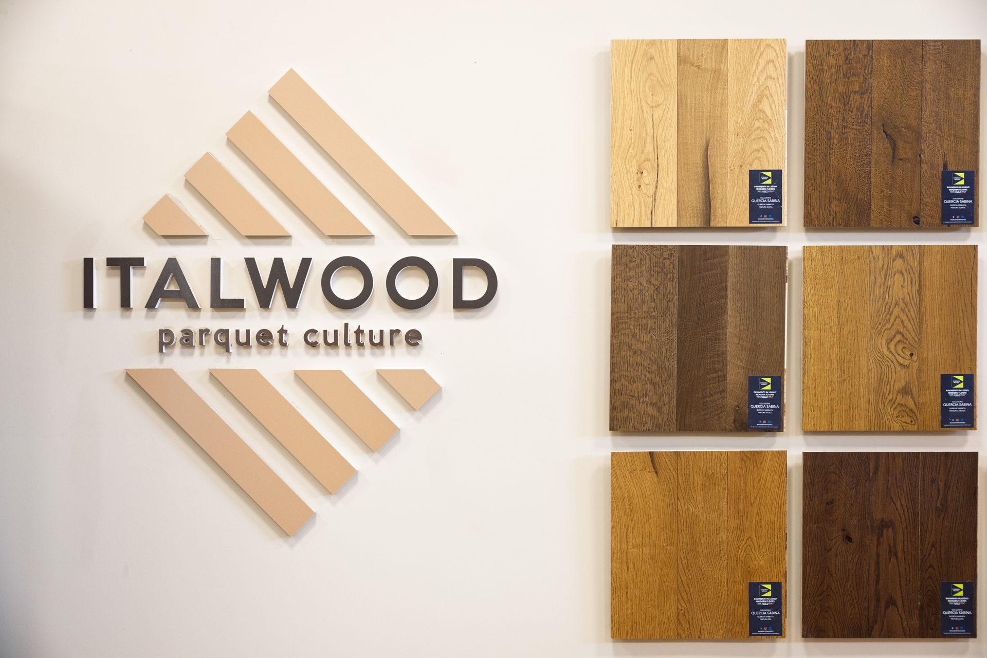 Il logo ITALWOOD su una parete accanto a sei campioni di pavimenti in legno esposti in uno showroom.