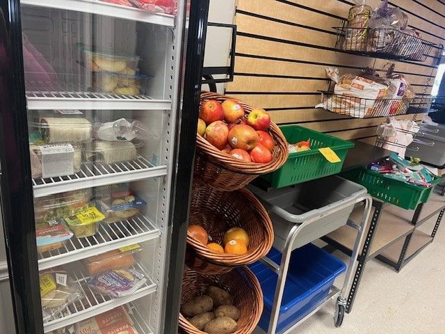 A refrigerator beside a tiered display of fresh apples, potatoes, and various packaged snacks on shelves and in bins.