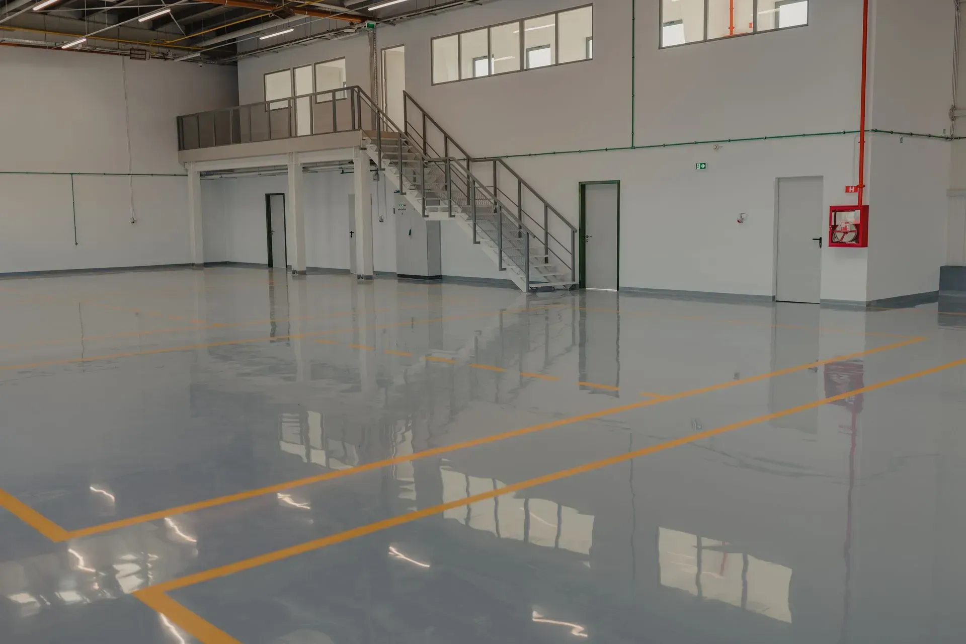 Spacious, gray-floored industrial interior. White walls, elevated office space with stairs, yellow floor markings, and fire equipment.
