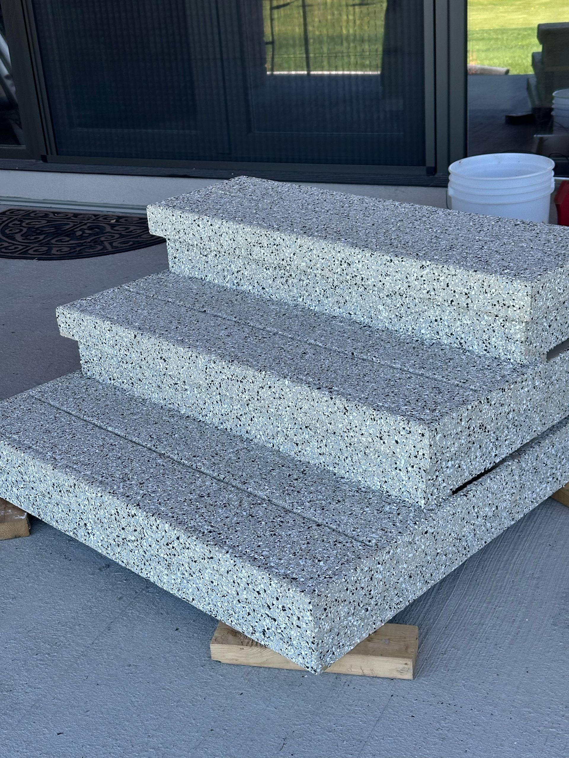Three stacked speckled stone steps resting on wooden blocks outdoors near a doorway