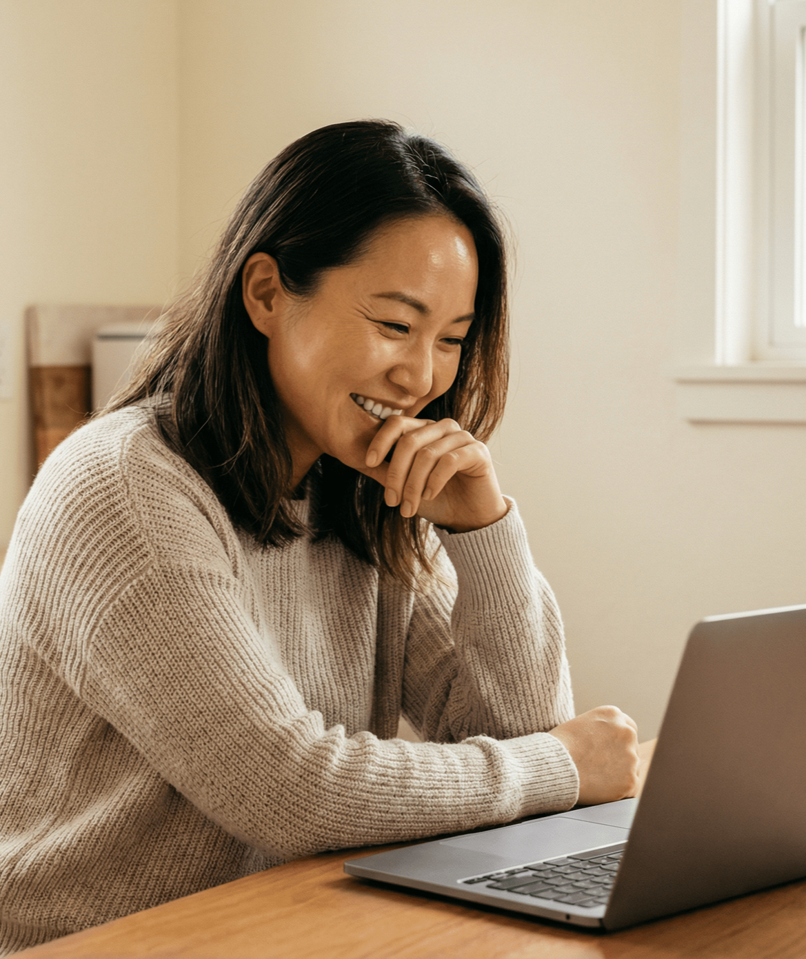 Woman smiling, looking at laptop on wooden table near window.