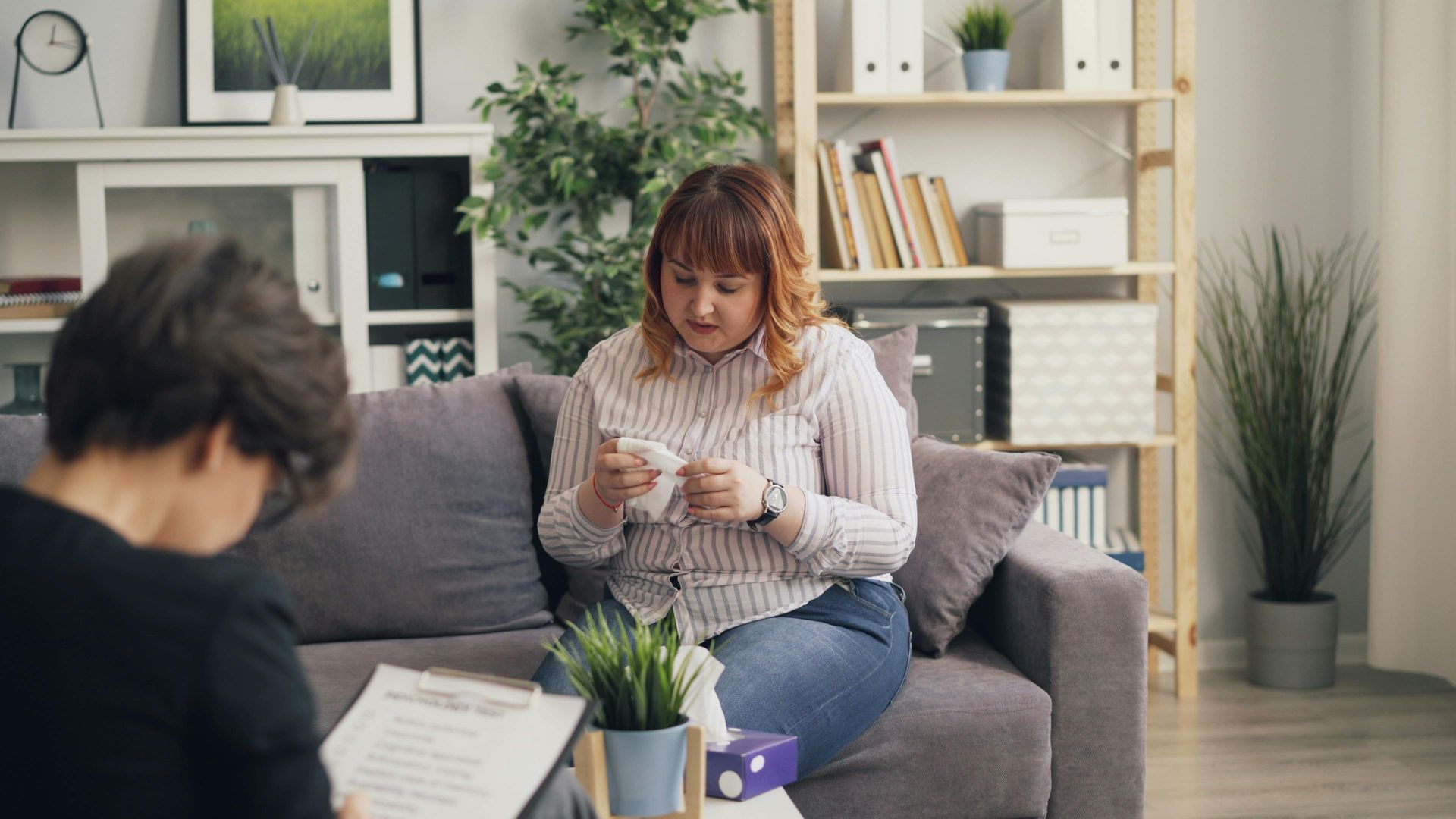 Woman knitting on a gray sofa in a bright living room, with another person blurred in the foreground.