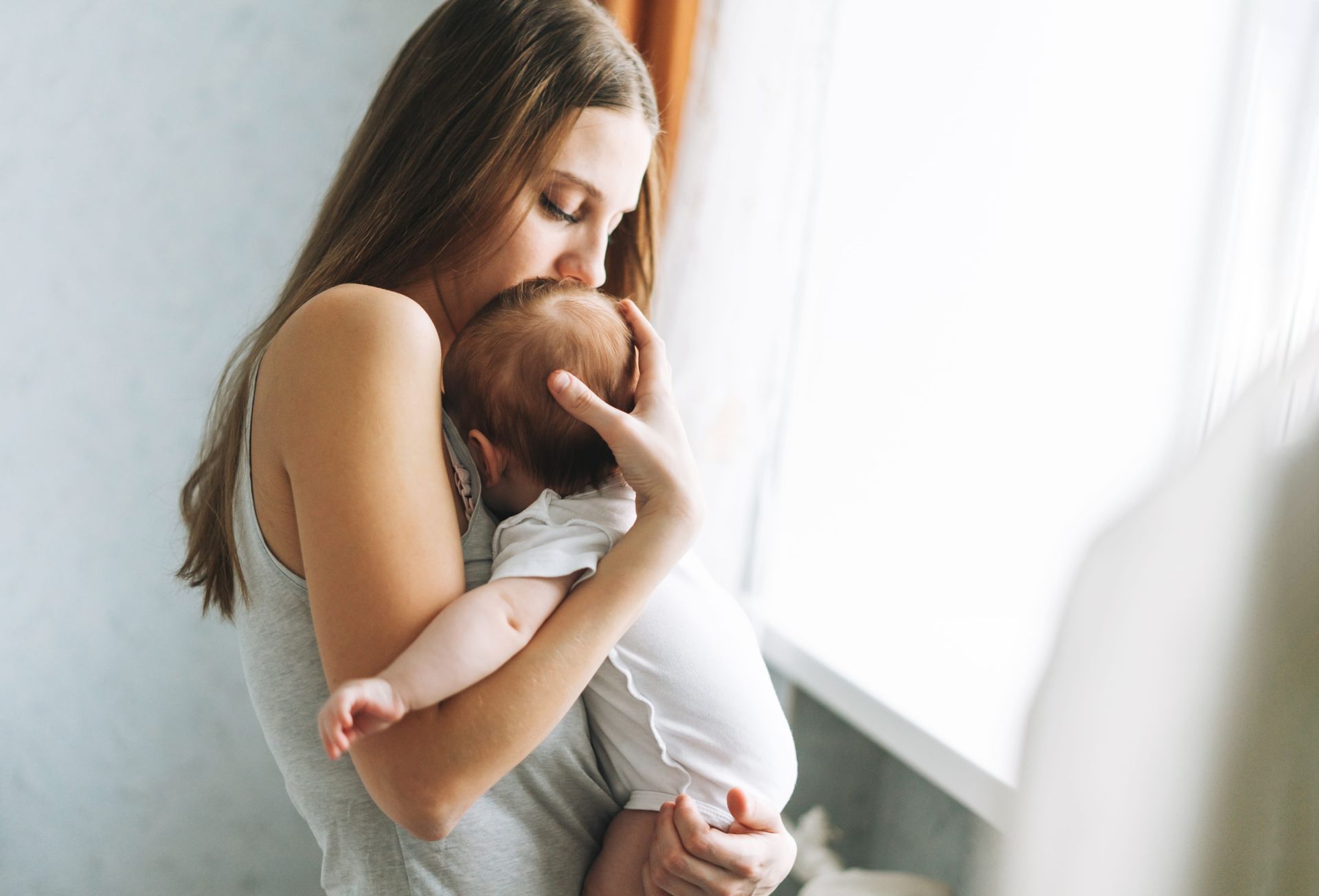 Woman holding and kissing a baby near a window.