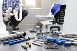 Plumber repairing bathroom pipes beside a toilet, with tools scattered on the floor.