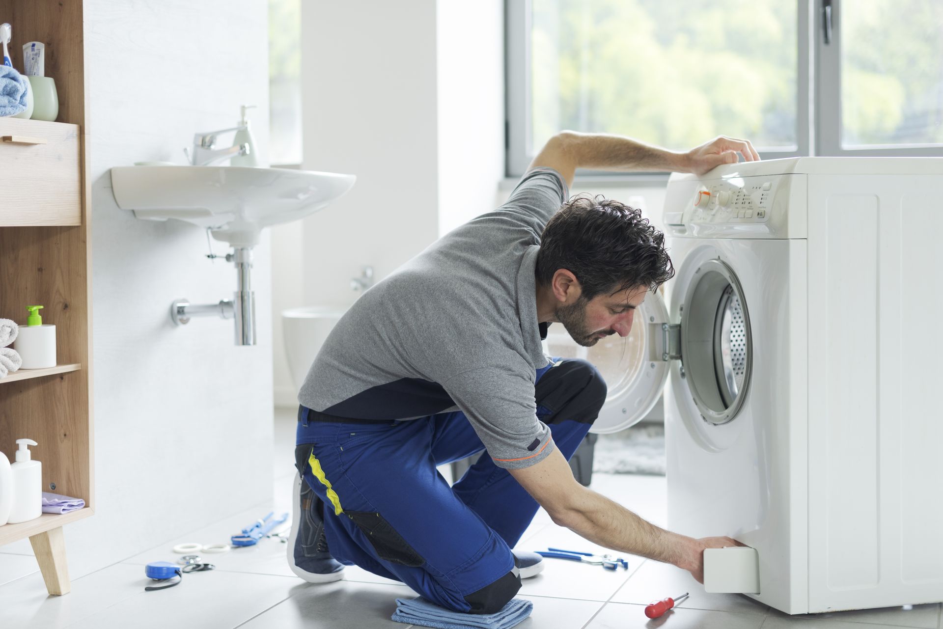 Man kneeling to repair a white washing machine in a bright laundry room
