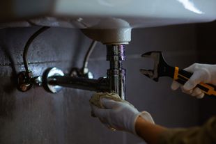 Plumber tightening pipes under a sink with a wrench and gloved hands