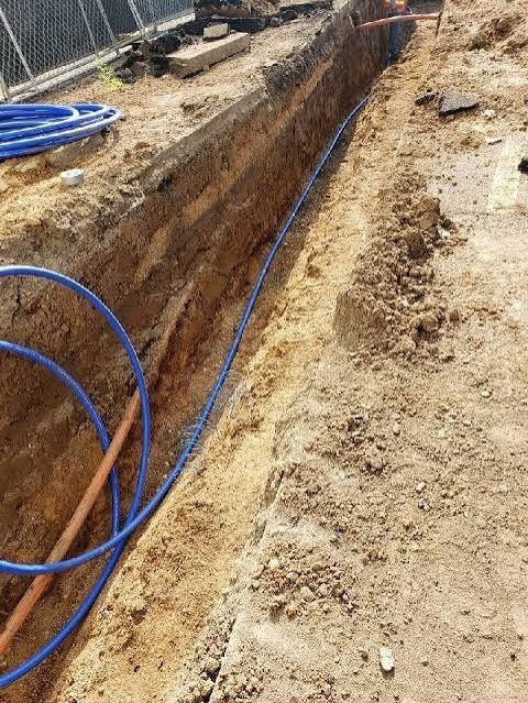 A narrow trench dug in sandy soil with blue utility pipes running along the bottom.