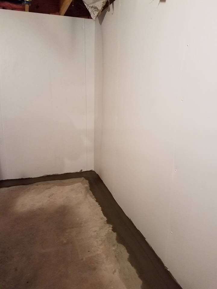 Corner of a basement with white walls and a concrete floor. A dark strip lines the floor where the walls meet.