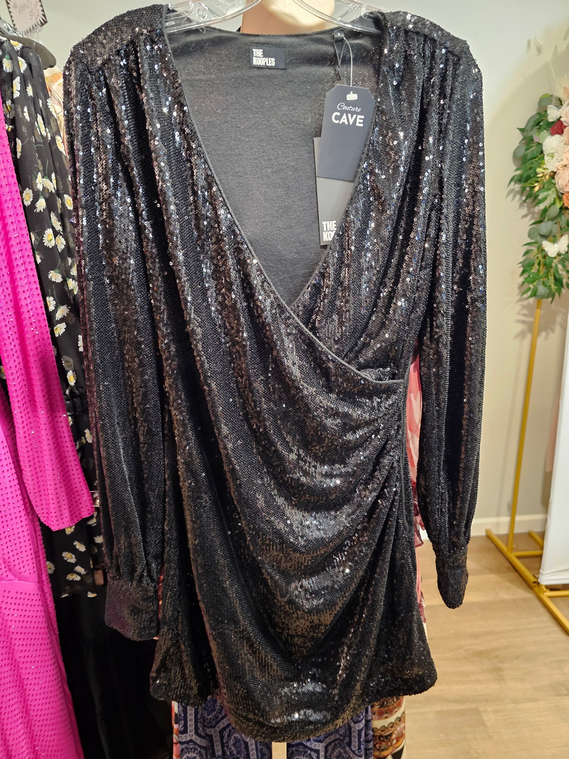 A black, long-sleeved, V-neck sequin mini dress with a draped, ruched front design hangs on a hanger in a store.
