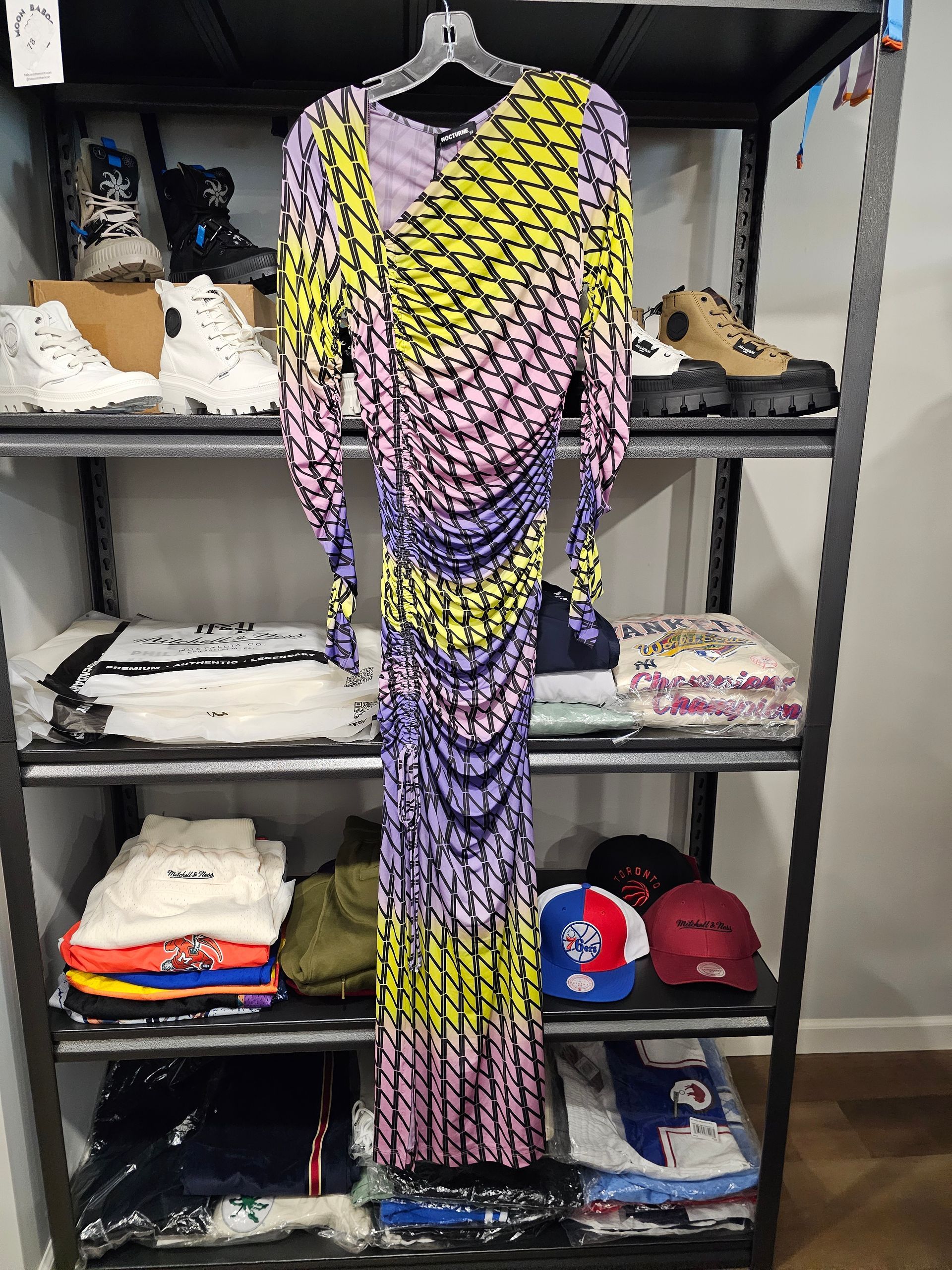 A long-sleeved, ruched midi dress with a diagonal yellow, purple, and black pattern hanging on a black wire shelf.