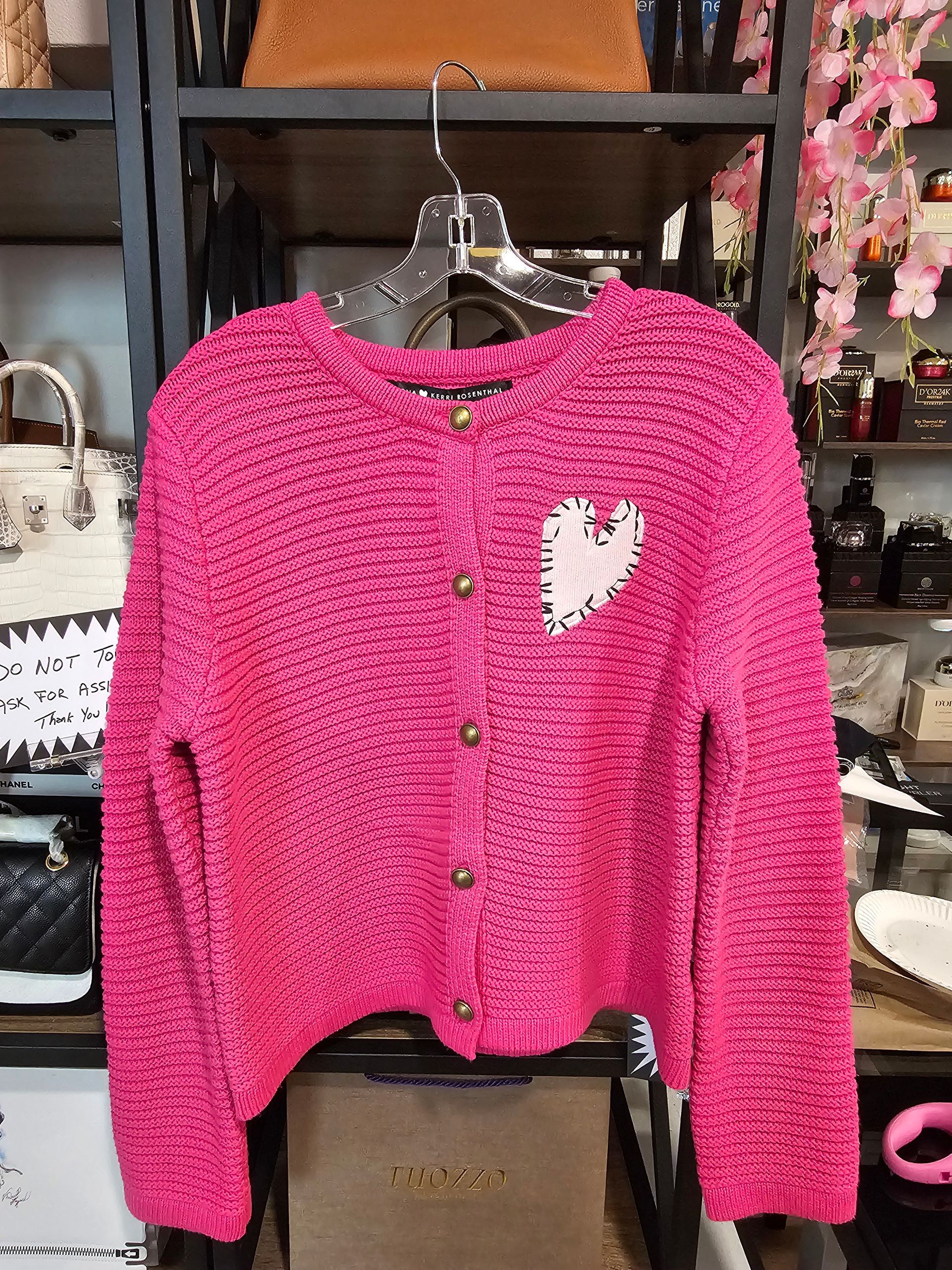 A bright pink, chunky knit cardigan with a small white heart patch on the chest, hanging on a hanger in a retail store.