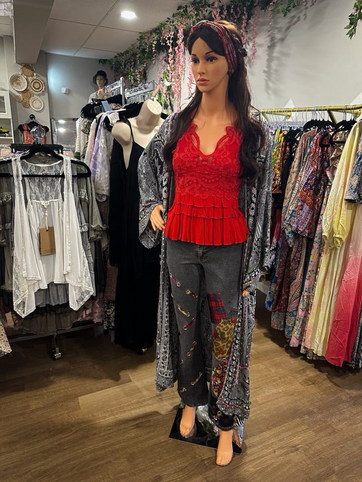 Mannequin in a boutique wearing red top, jeans, and patterned coat. Clothing racks and shelves filled with garments.