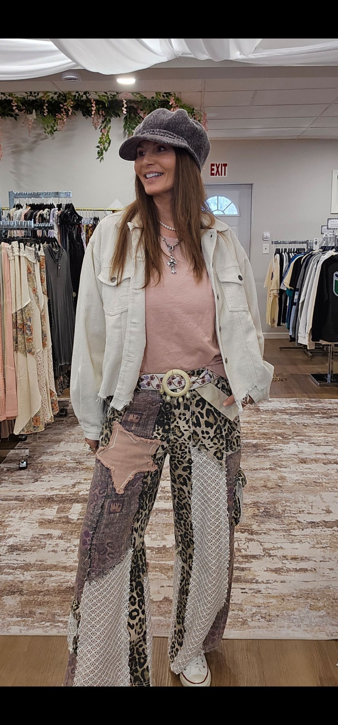 Woman in a store wearing a hat, jacket, pink shirt, and patterned pants. She is smiling.
