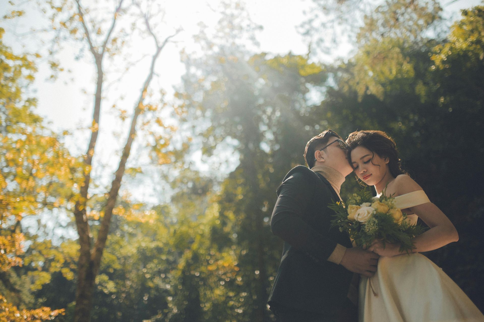 Couple embracing outdoors; the man kisses the woman's cheek, holding flowers, sunlight.