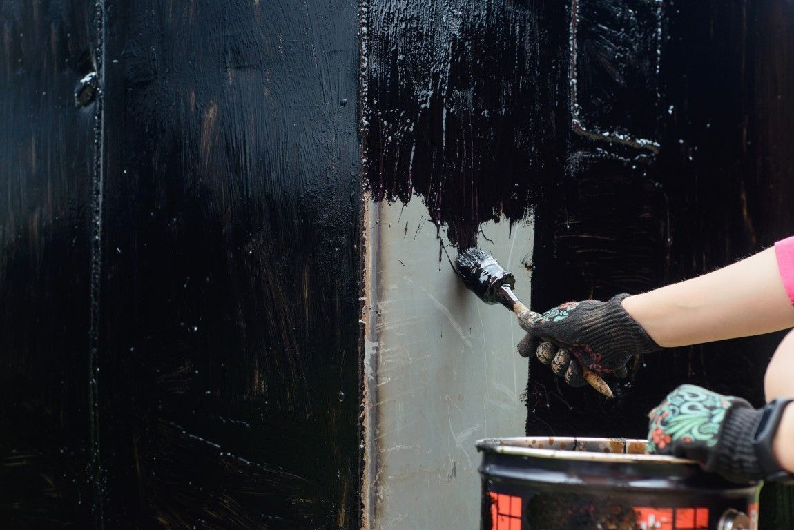 Gloved hand painting a black wall with a roller, covering a gray patch beside a paint bucket