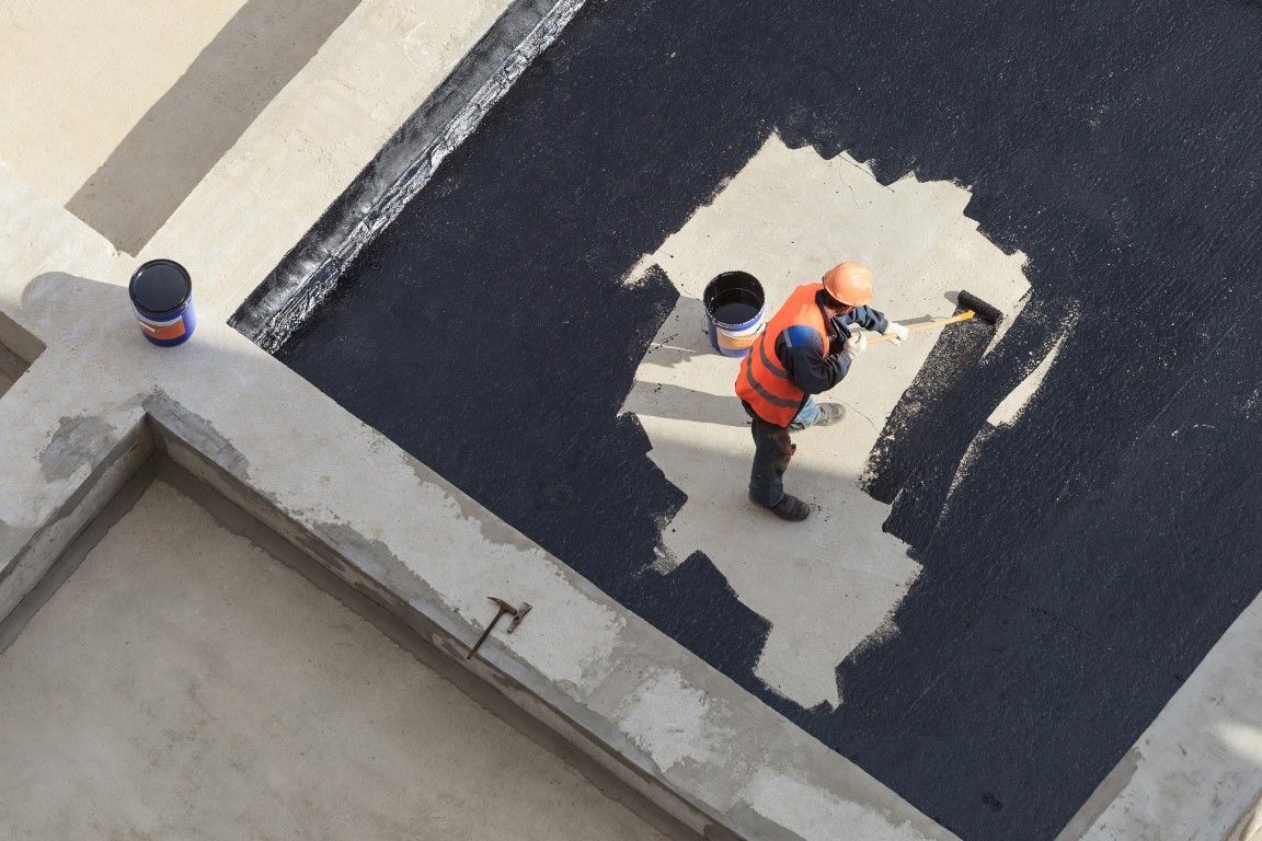 Worker in orange vest painting a dark rooftop, with a lighter patch of fresh coating.