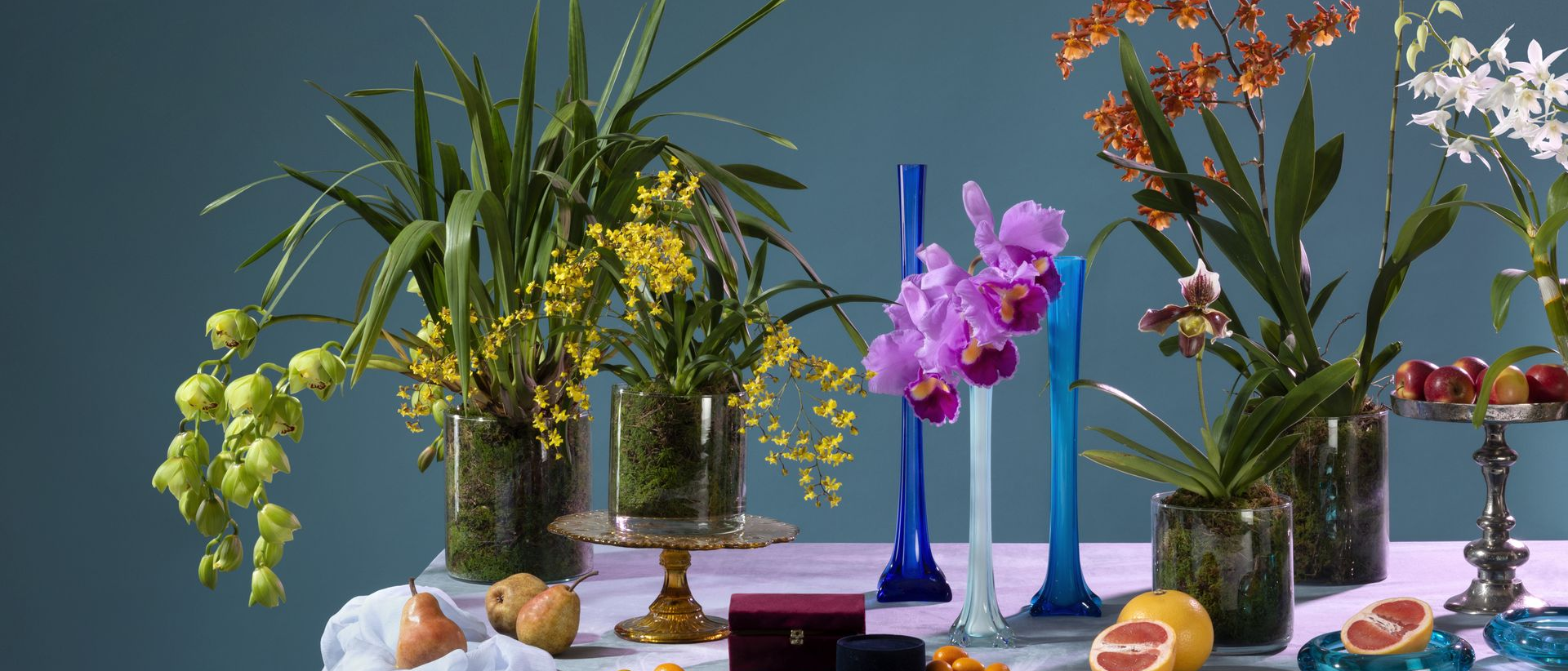 Colorate composizioni floreali in vasi, con candele e frutta, su un tavolo davanti a uno sfondo blu.