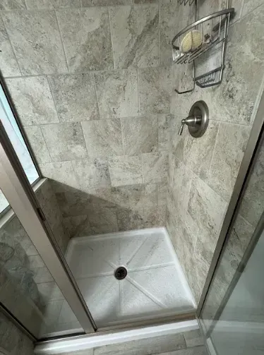 Shower stall with gray tiled walls, white shower base, and sliding glass doors. Soap holder and showerhead present.