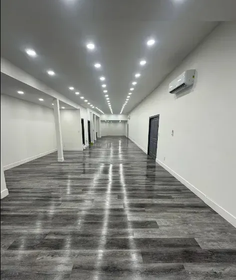 Empty, long room with gray wood-look flooring, white walls, and bright overhead lights.