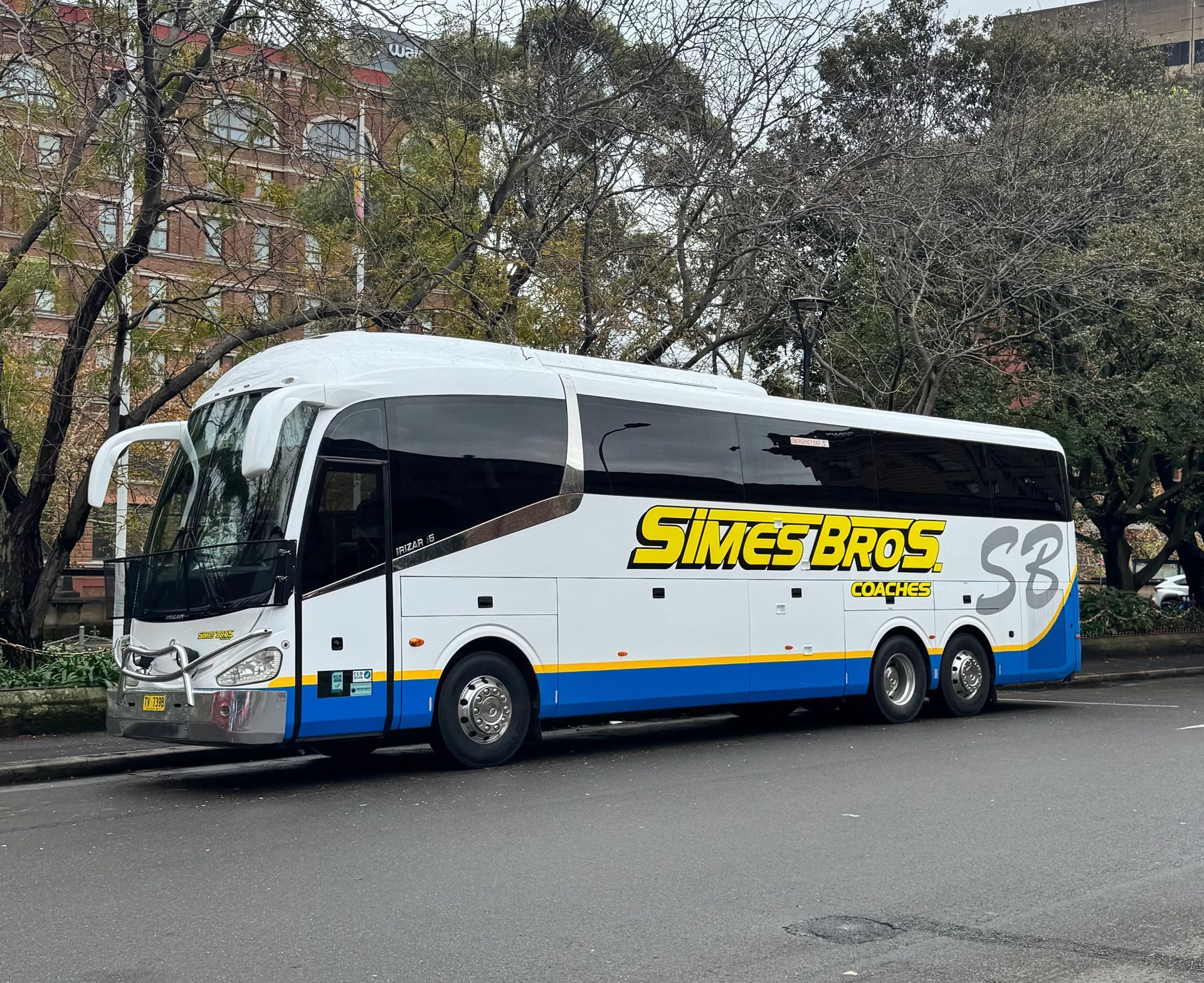 White and blue Simes Bros. coach bus parked on a street with a brick building in the background.