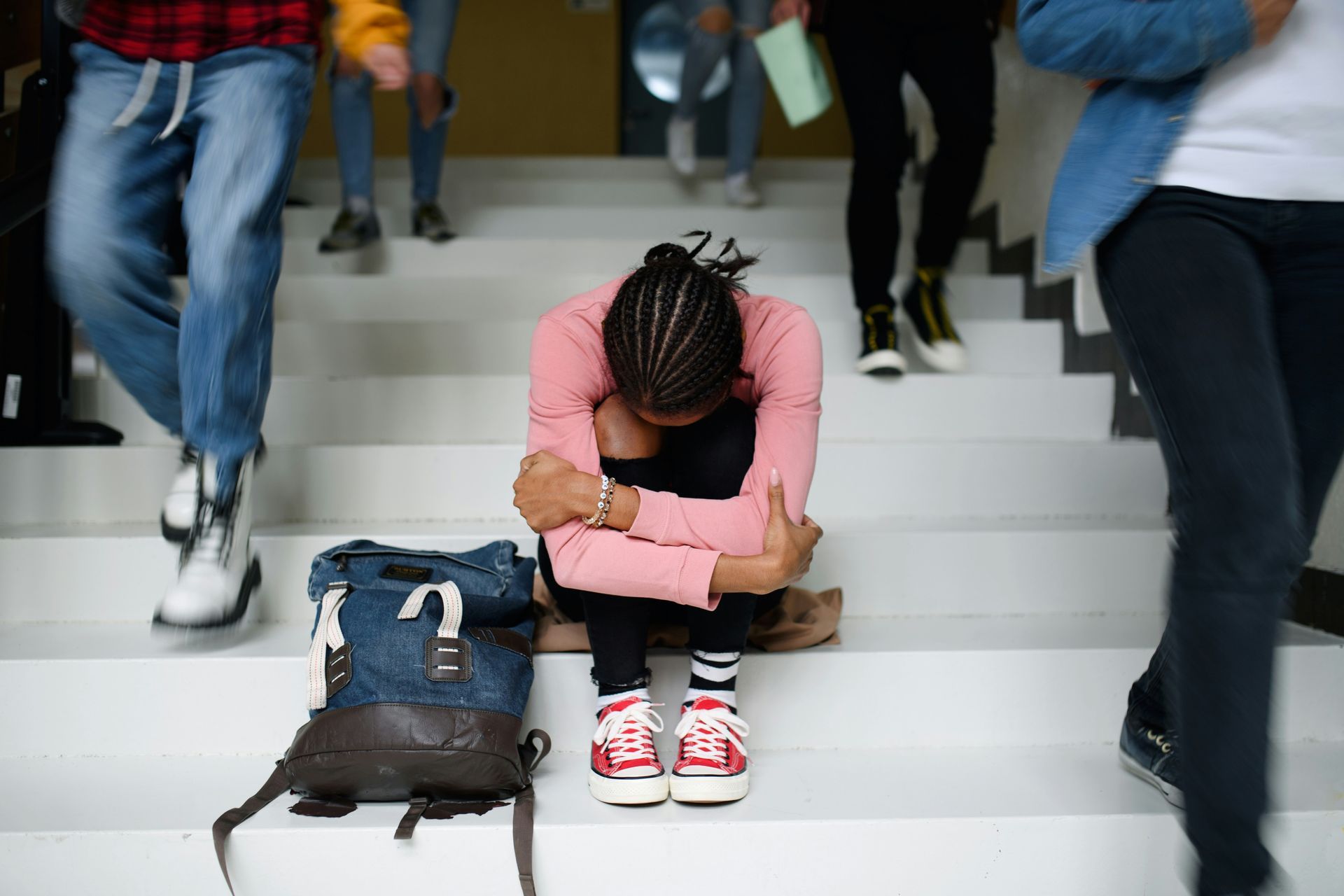 A student sits on school steps with her head down and alone as other classmates walk by them.