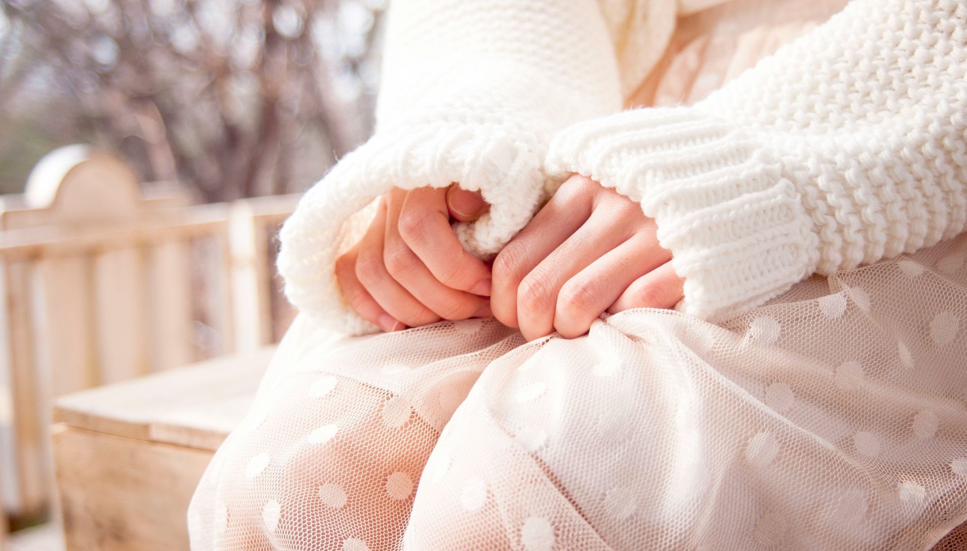 Child in a white sweater sitting outdoors with hands folded in their lap.