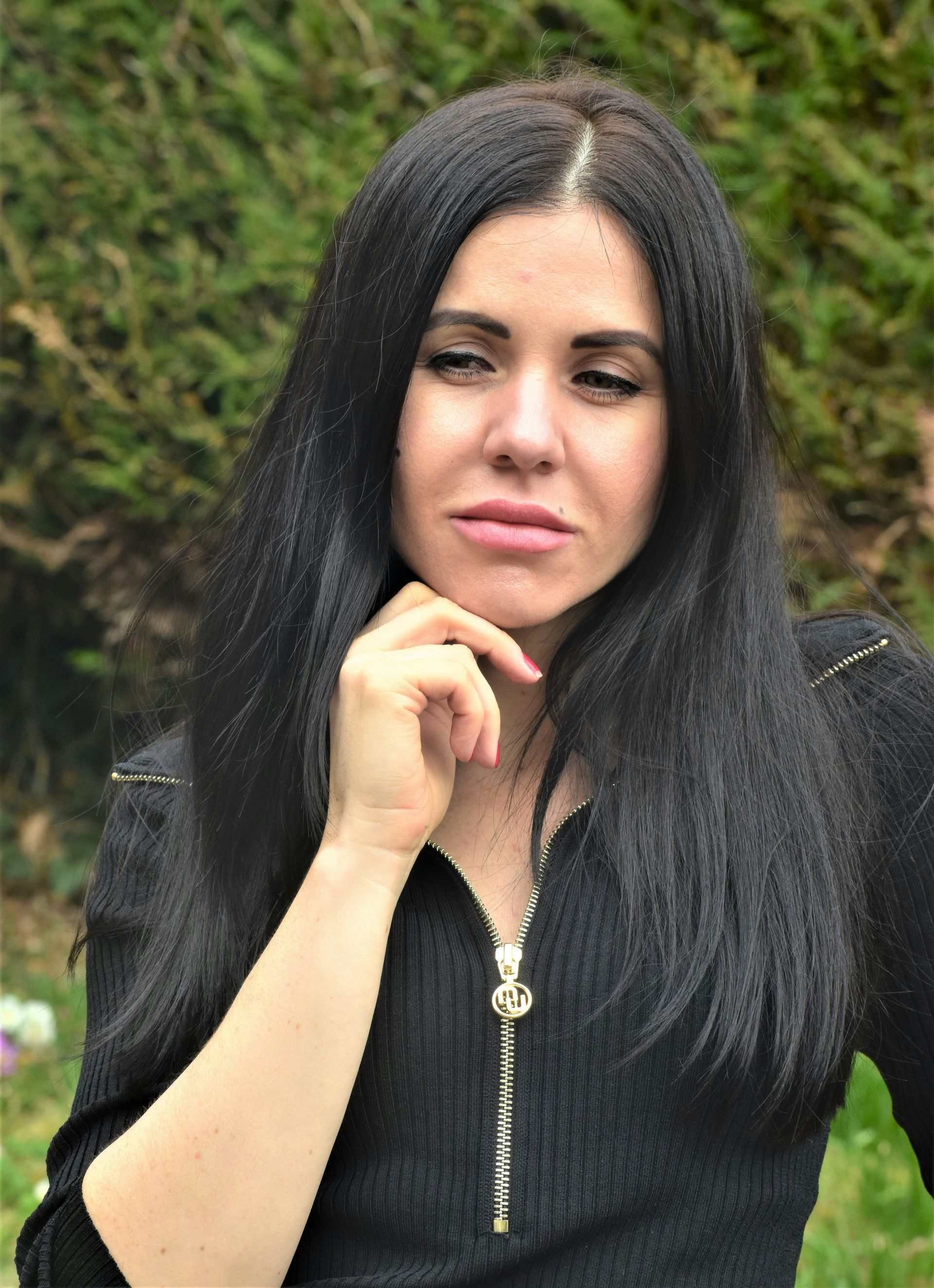 A woman with long dark hair, looking thoughtful, outdoors with greenery in the background.